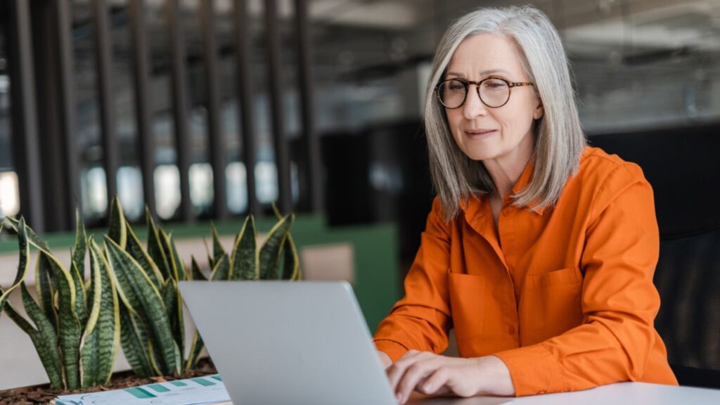 Confident serious mature businesswoman wearing stylish eyeglasses, orange shirt using laptop working online in modern office. Manager planning project, typing on keyboard. Work Concept