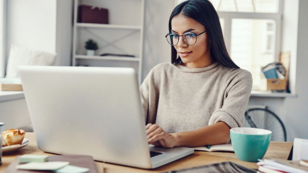 Beautiful young woman in casual clothing using laptop and smiling while working indoors. Work Concept