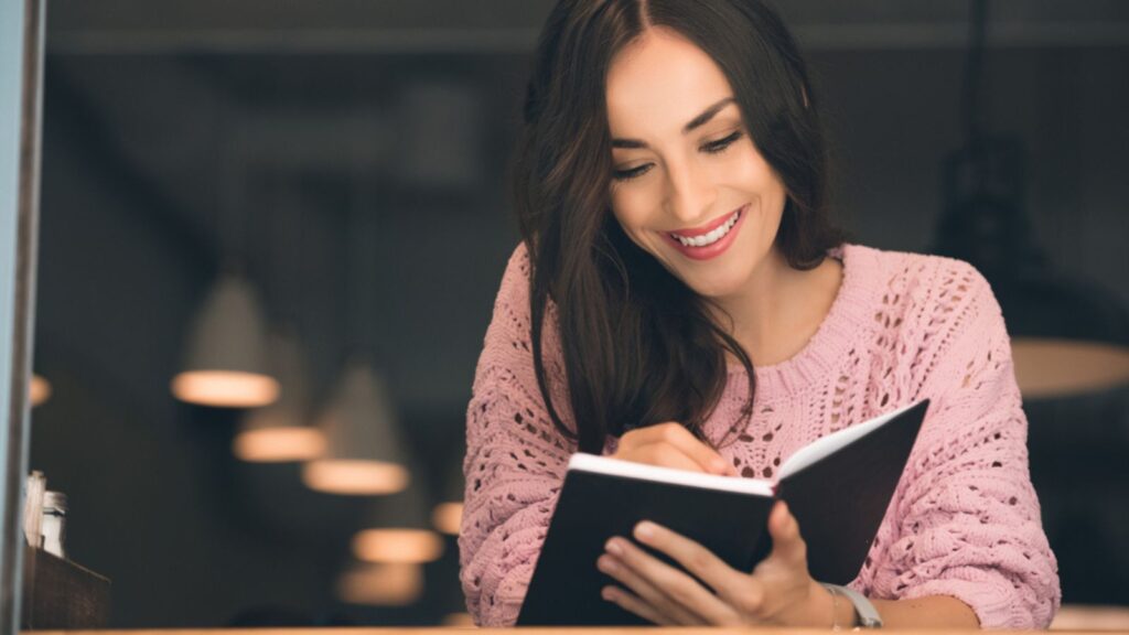 Happy young woman making notes in textbook at table in cafe