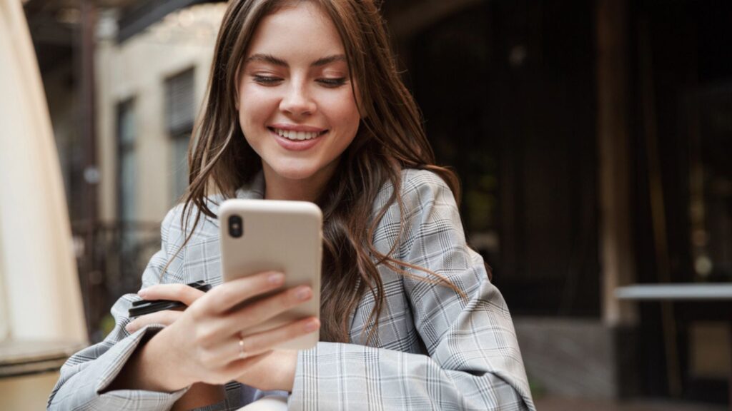 Beautiful young woman using phone while sitting at the cafe table outdoors