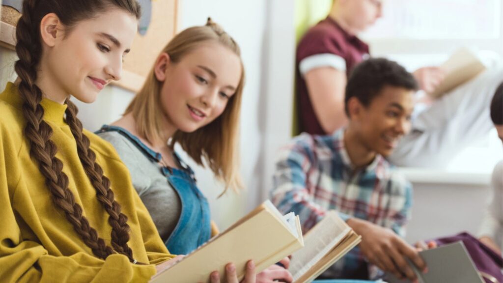 Group of smiling multicultural teenage schoolchildren reading books during school break