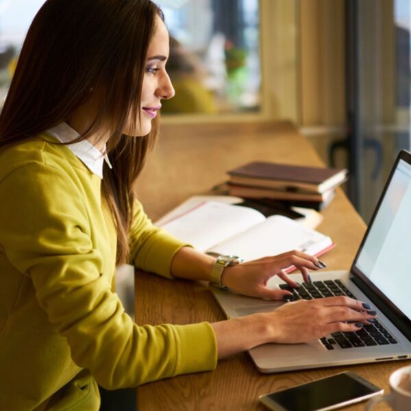 Positive young female journalist sitting with pen in hand at table with markers notebook and working on laptop while looking at screen during online information gathering for publication in cafe
