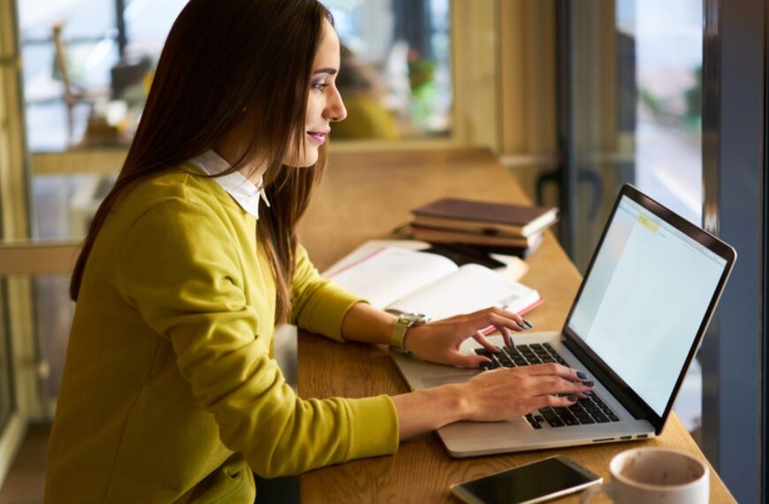 Positive young female journalist sitting with pen in hand at table with markers notebook and working on laptop while looking at screen during online information gathering for publication in cafe