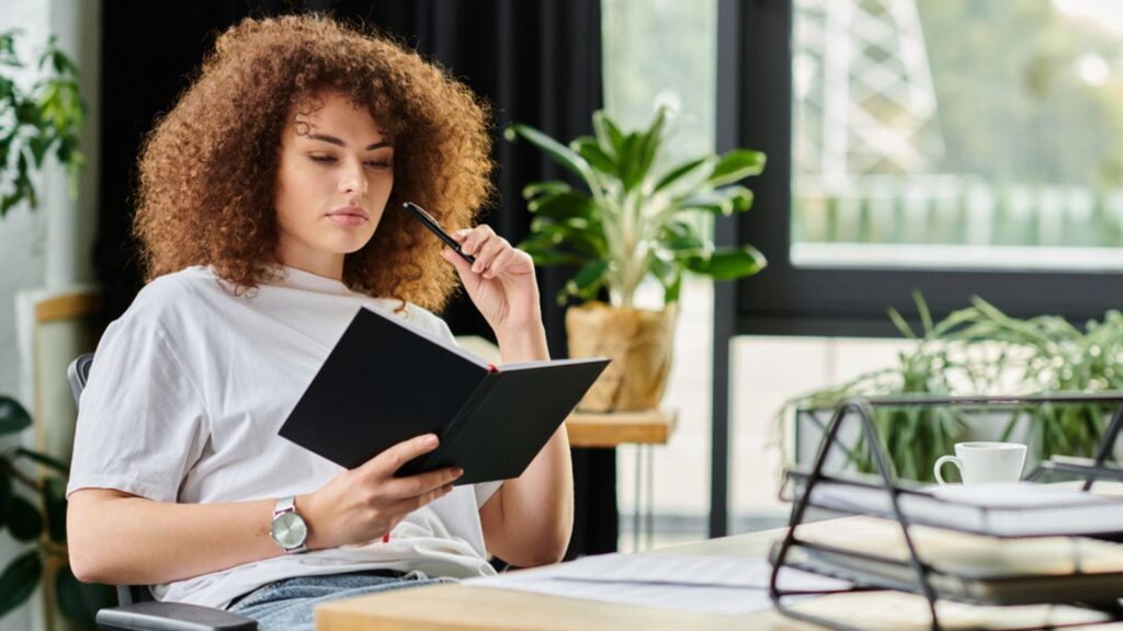 A woman sits quietly, pondering and writing her notes while surrounded by plants in a cozy workspace