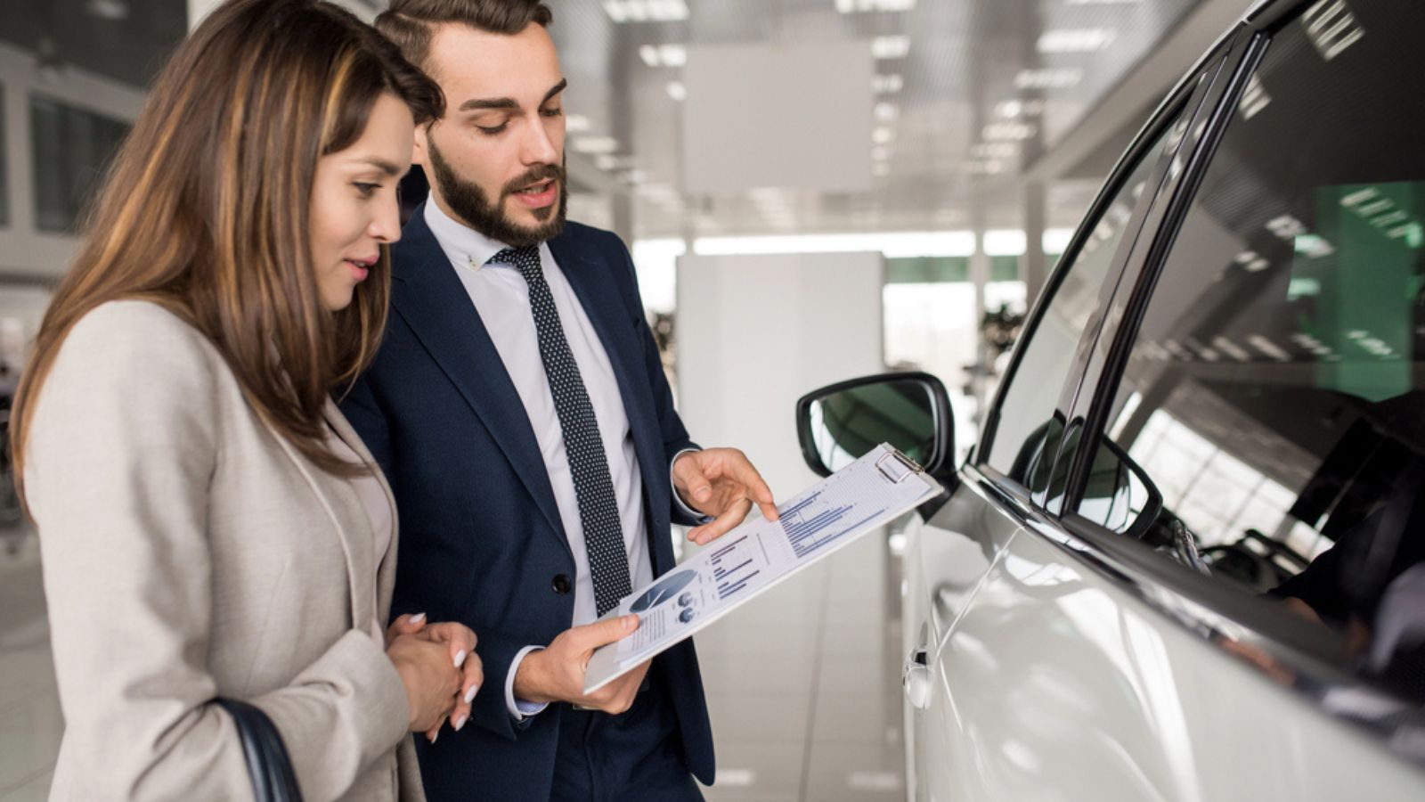 Side view portrait of modern young woman listening to car dealer while choosing luxury car in showroom