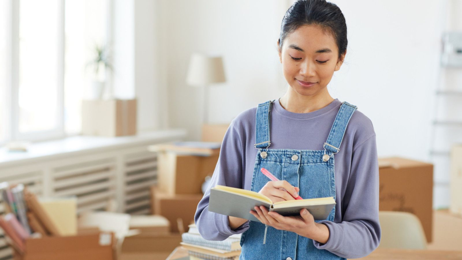 Waist up portrait of young Asian woman writing in planner while standing in new house or apartment with cardboard boxes in background, copy space. Home Organization.